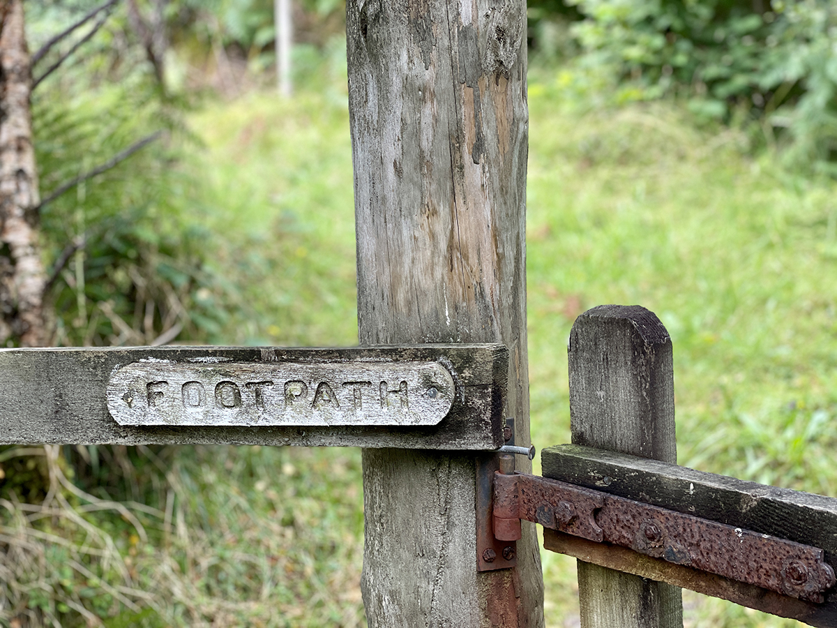 A wood post and rail fence with a rusty gate hinge and an engraved wooden sign that says FOOTPATH.