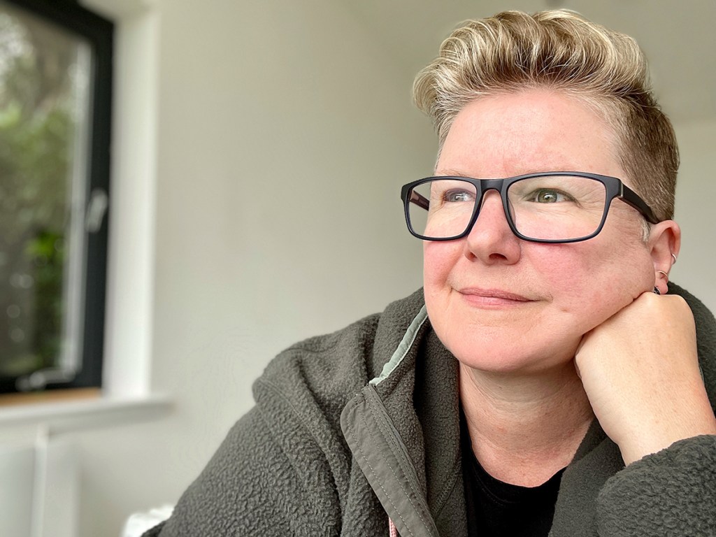 The artist - a woman with fair skin and very short fair hair in a khaki green fleece and wearing dark blue rimmed glasses staring out a window in the studio.